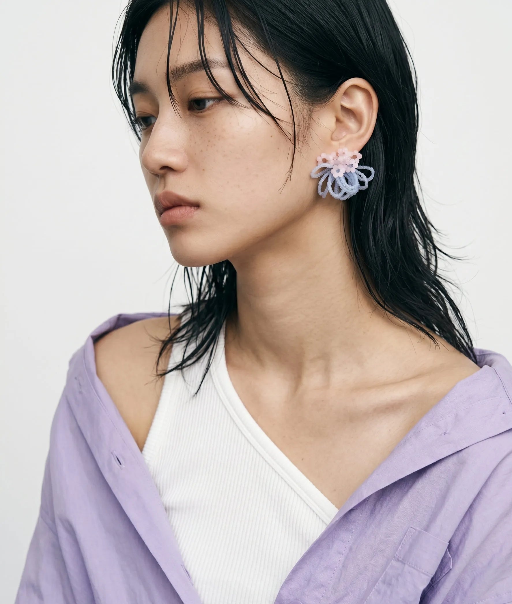 A close-up studio portrait of a woman showcasing premium effortless K-style with dewy makeup, natural freckles, and trendy wet-look hair. She wears a casually draped lavender shirt over an asymmetrical white top, perfectly highlighting unique handcrafted statement beaded cluster earrings with pastel pink and light blue accents.
