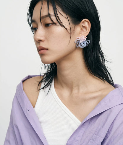 A close-up studio portrait of a woman showcasing premium effortless K-style with dewy makeup, natural freckles, and trendy wet-look hair. She wears a casually draped lavender shirt over an asymmetrical white top, perfectly highlighting unique handcrafted statement beaded cluster earrings with pastel pink and light blue accents.