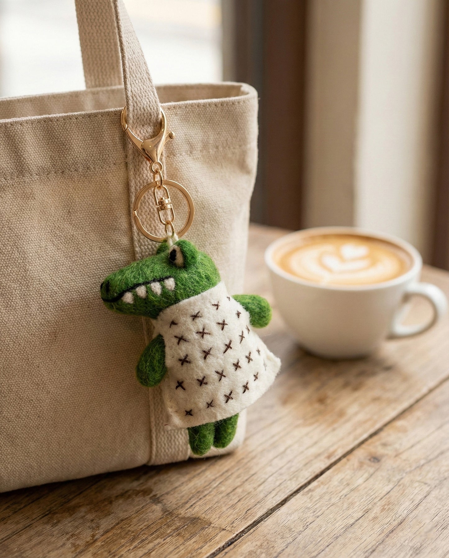 Green plush toy with a patterned dress attached to a beige bag on a wooden surface with a cup of coffee.