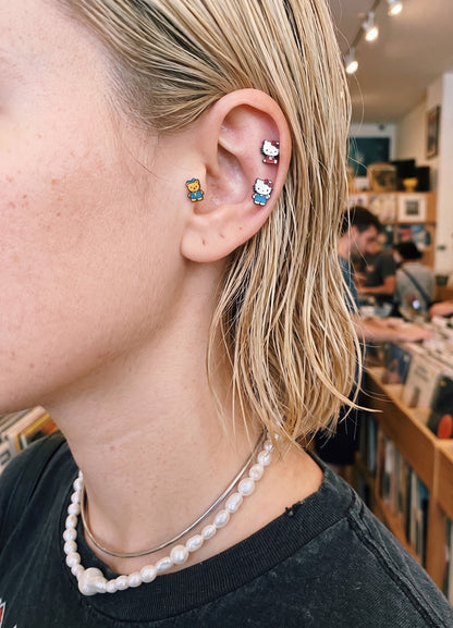 Ear-terior close-up — Hello Kitty Studs, Tiny Chum and Rilakkuma character piercing stacked on helix and conch, Kidcore Y2K jewelry with pearl necklac
