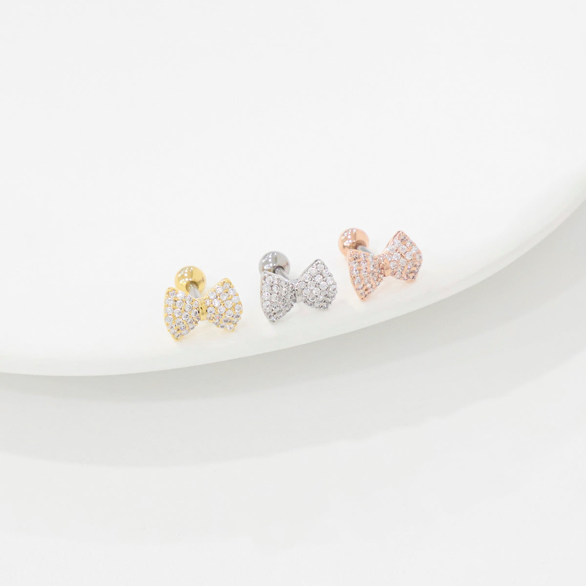 Gold, silver, and rose gold bow barbell earrings aligned diagonally on a white plate with soft studio lighting
