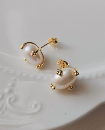 Gold pearl stud earrings in an oval shape resting on a white ceramic plate.