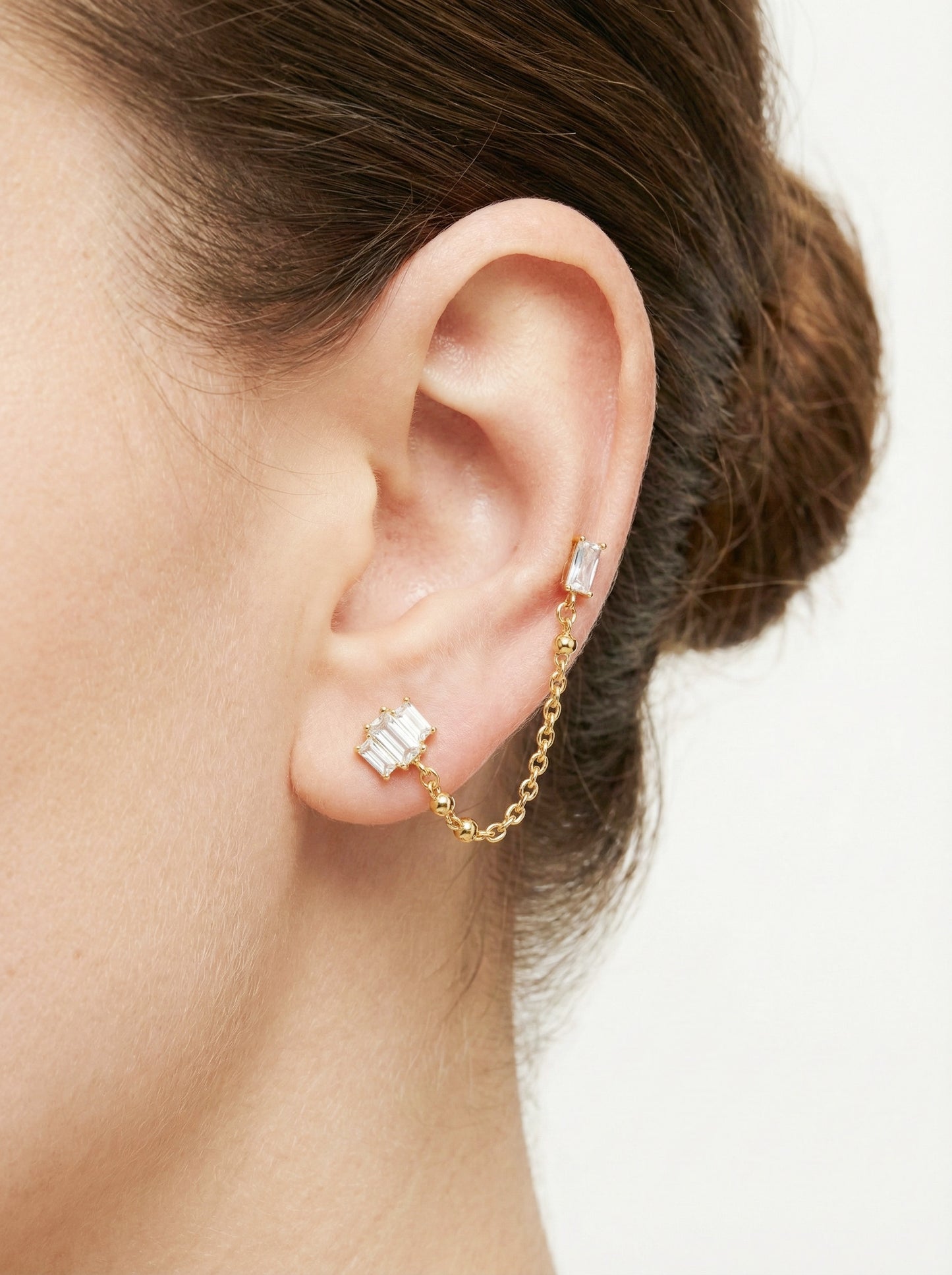 A woman's ear adorned with a gold Baguette CZ Chain Double Stud Earring, featuring baguette-cut cubic zirconia linked by a gold chain, surgical steel base.