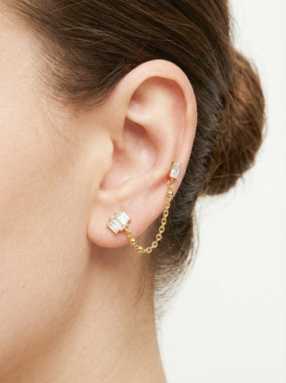 A woman's ear adorned with a gold Baguette CZ Chain Double Stud Earring, featuring baguette-cut cubic zirconia linked by a gold chain, surgical steel base.