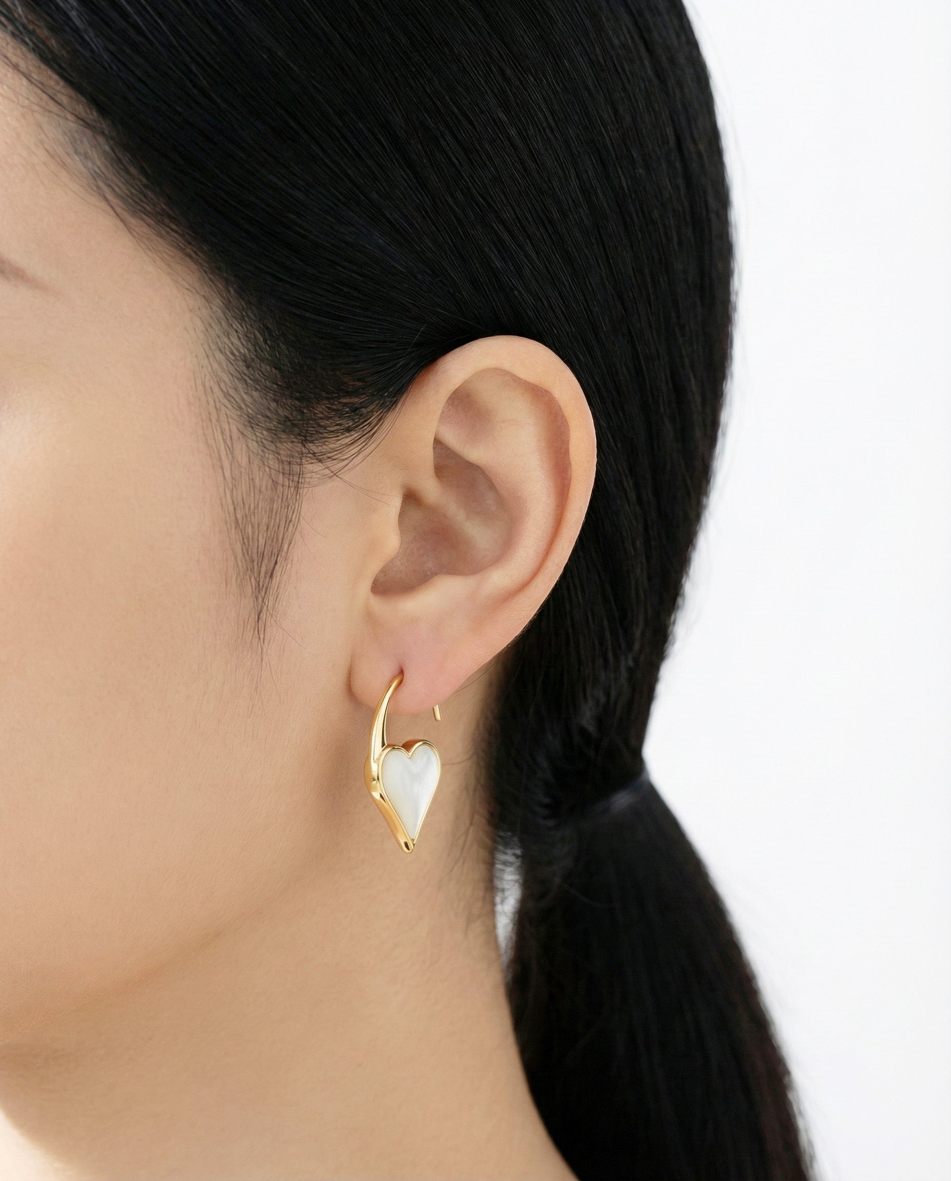 Gold heart hook earring with a white mother-of-pearl inlay, worn by a woman with a low ponytail against a soft white background.