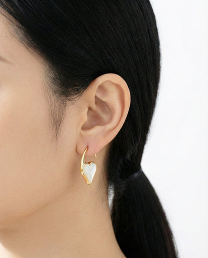 Gold heart hook earring with a white mother-of-pearl inlay, worn by a woman with a low ponytail against a soft white background.