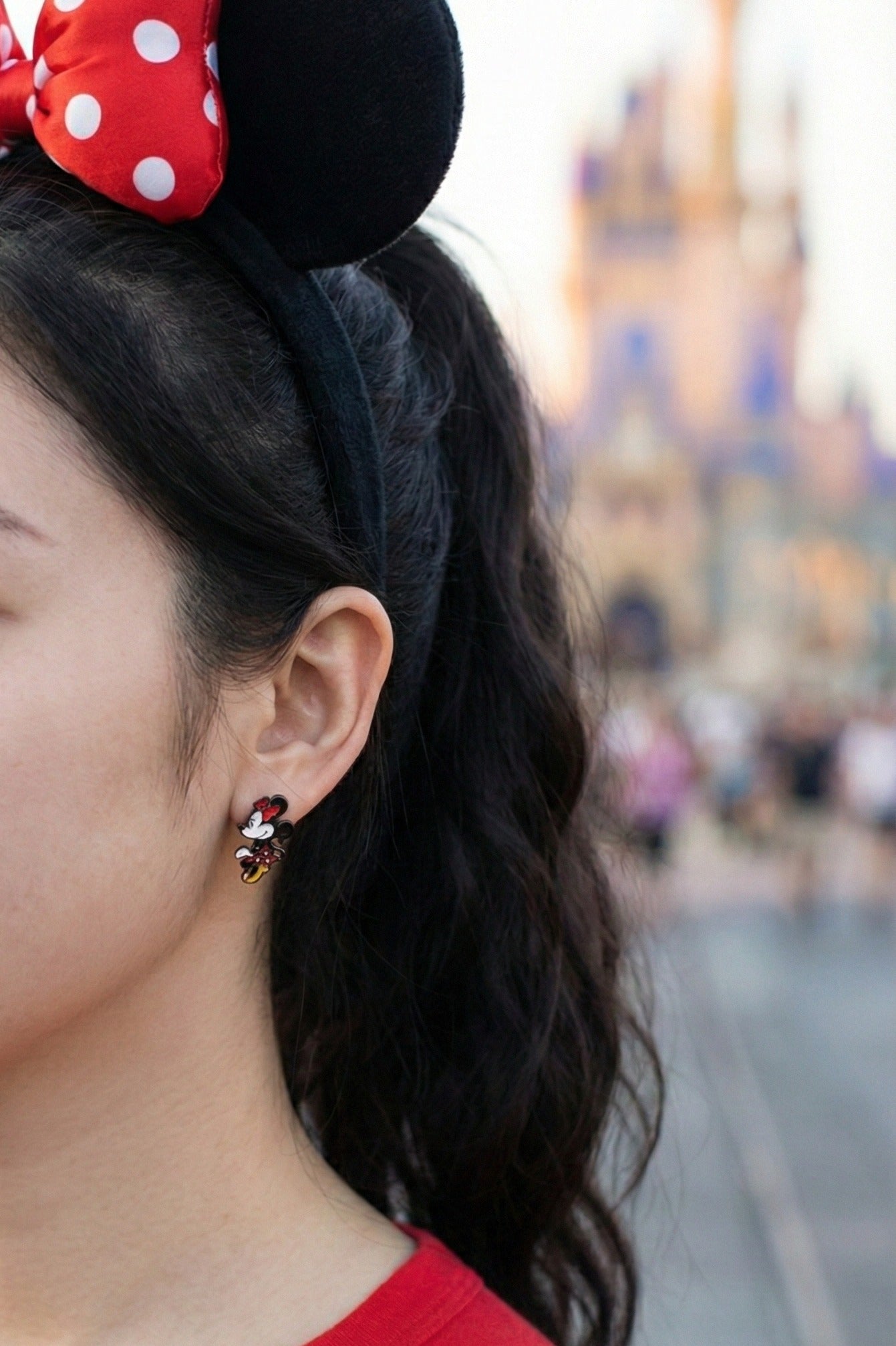 Woman wearing Disney Minnie Mouse front-back 3D earrings at Disneyland castle with red polka dot headband