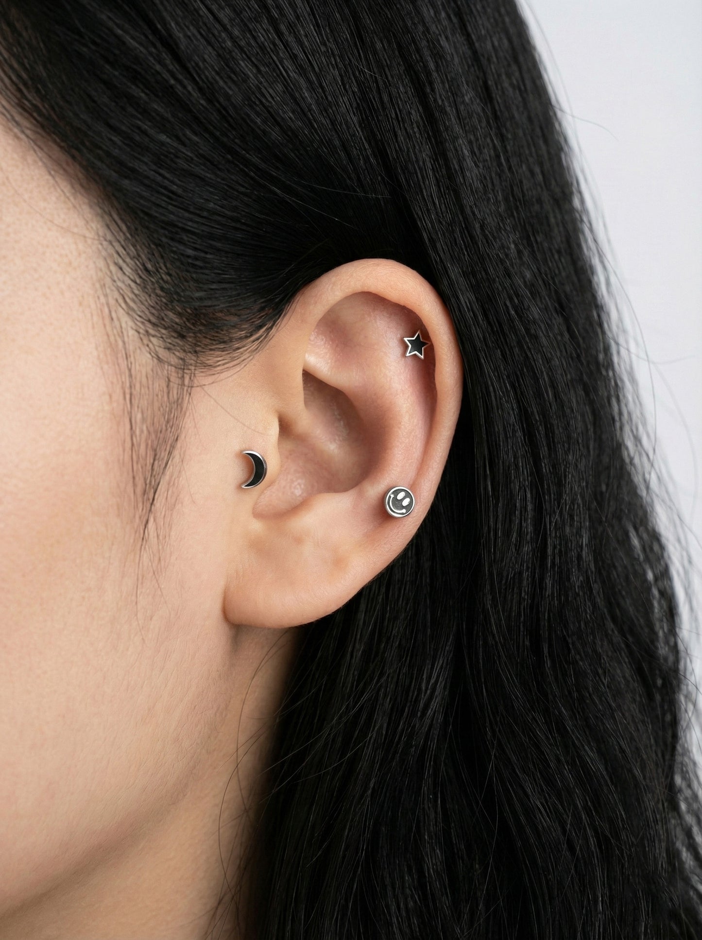  Close-up ear piercing stack with black moon, smiley stud, and silver star on cartilage