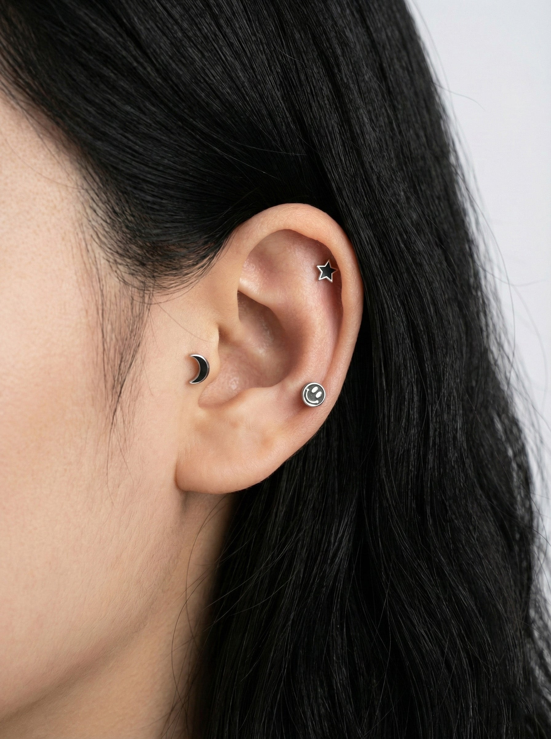  Close-up ear piercing stack with black moon, smiley stud, and silver star on cartilage