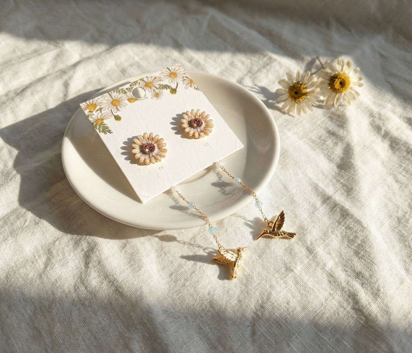 Close-up of gold daisy stud earrings with hummingbird charm on earring card