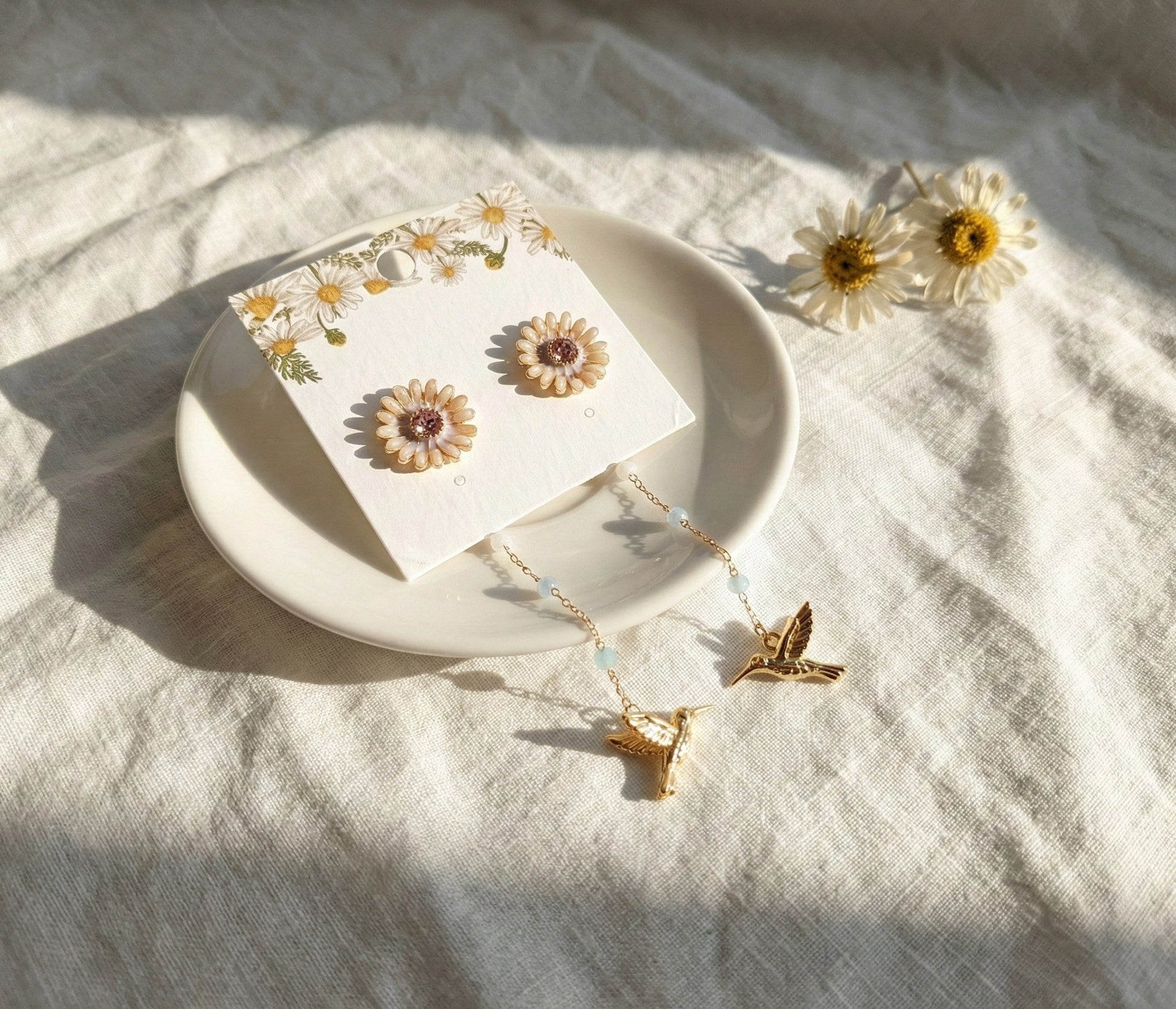 Close-up of gold daisy stud earrings with hummingbird charm on earring card