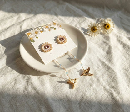 Close-up of gold daisy stud earrings with hummingbird charm on earring card