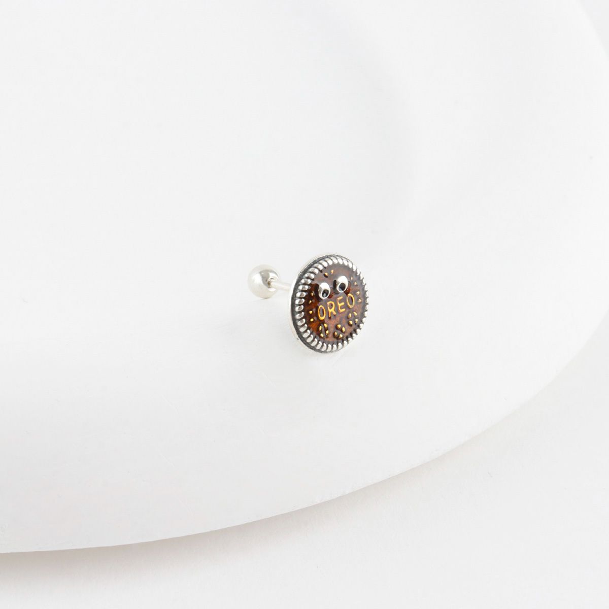 Close-up of brown Oreo cookie surgical steel stud piercing with 925 silver post, detailed texture and googly eyes on white background