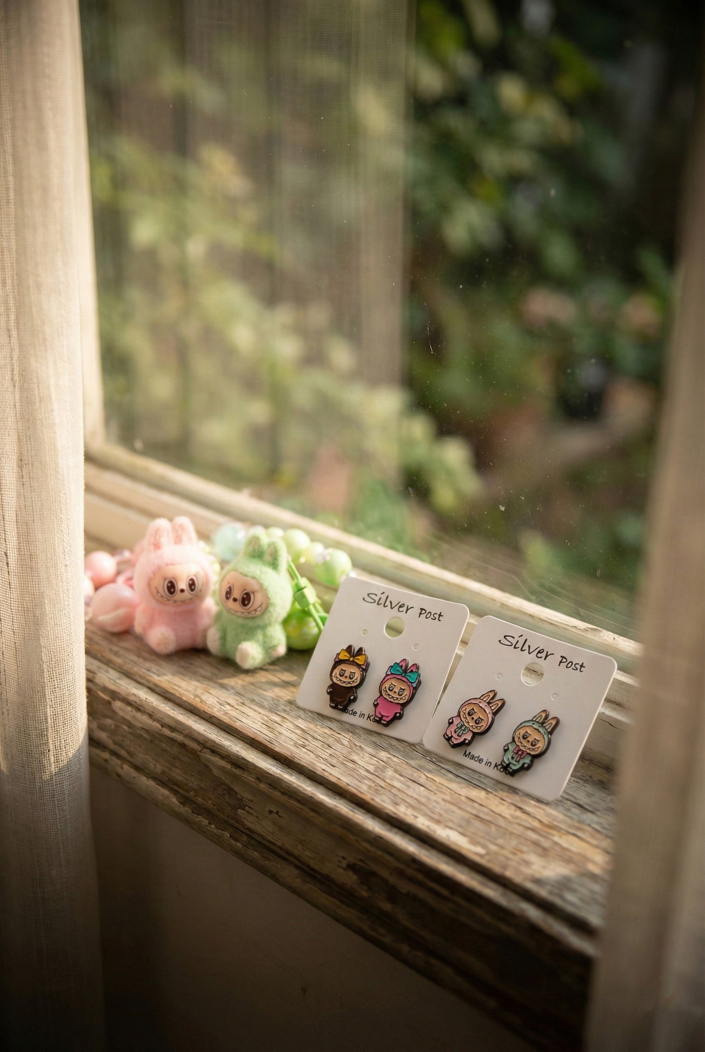 White earring cards with Labubu pink and brown bunny earrings on wooden windowsill with pink and green Labubu plush toys