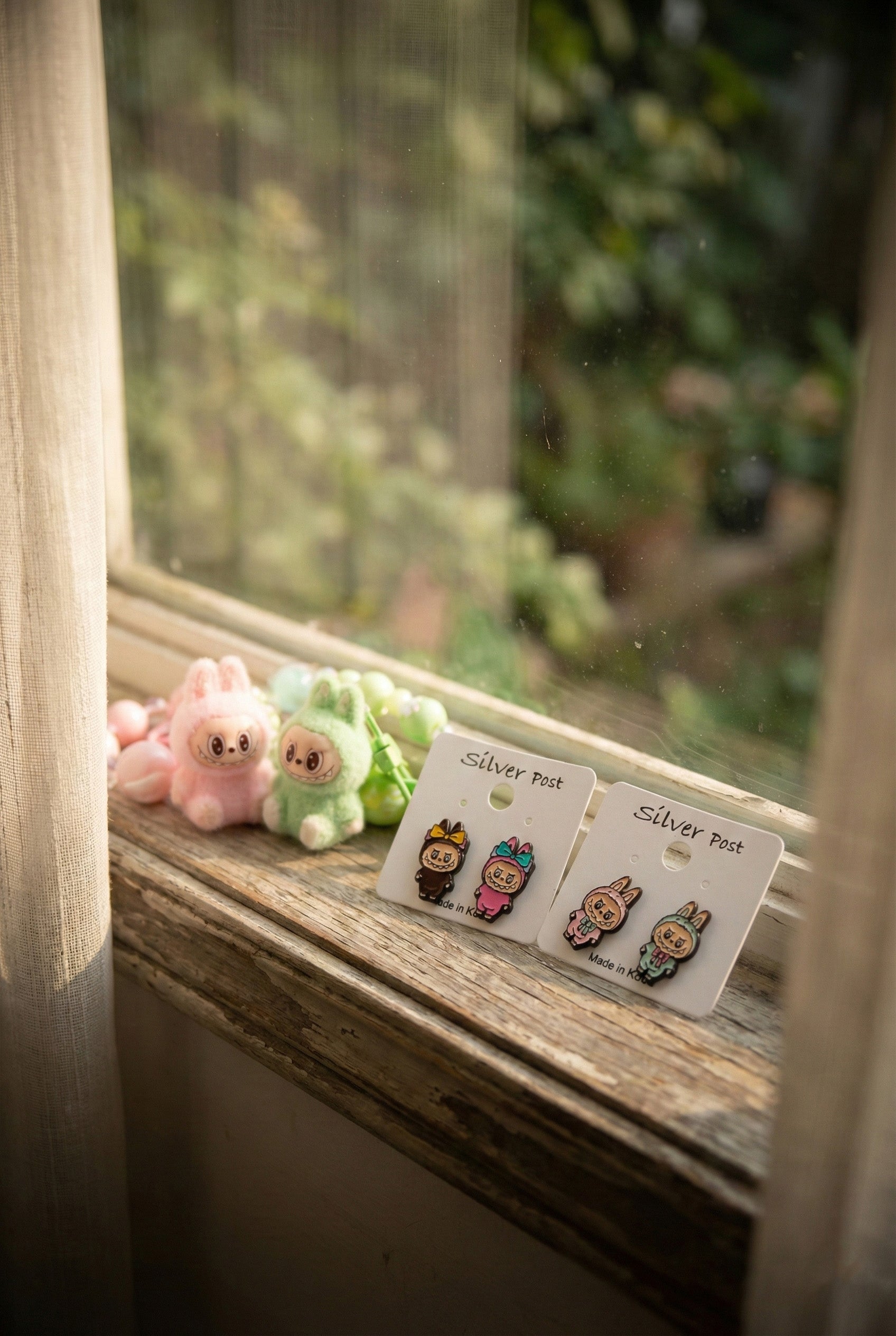 White earring cards with Labubu pink and brown bunny earrings on wooden windowsill with pink and green Labubu plush toys