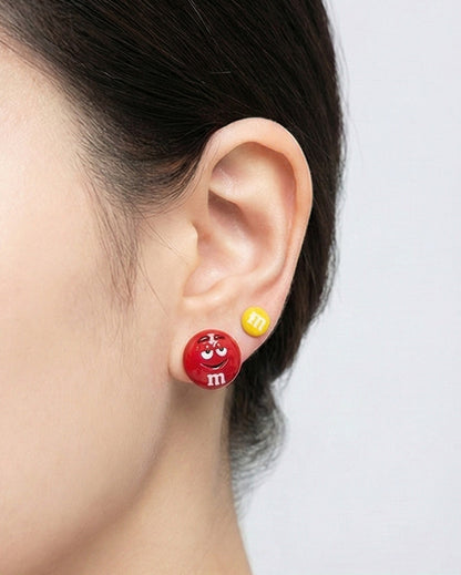 Side view of woman wearing M&M's chocolate candy character earrings, red smiling face with yellow mini stud in double lobe piercing, enamel coated brass with hypoallergenic silver posts, Korean kawaii jewelry