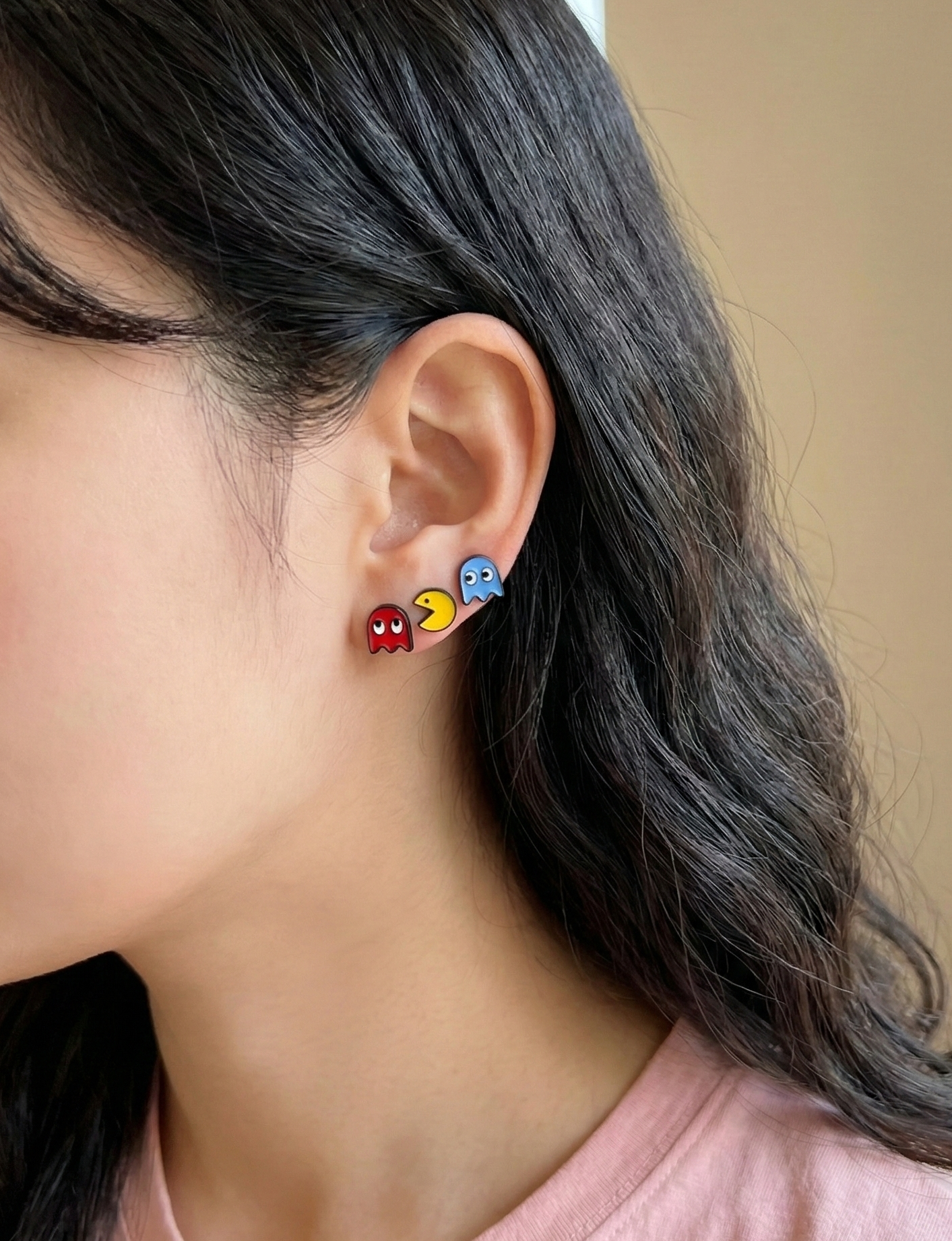 Woman wearing three layered stud earrings — red ghost, yellow Pac-Man, and blue ghost — stacked across two ear piercings for a retro arcade layered look