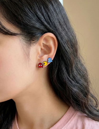 Woman wearing three layered stud earrings — red ghost, yellow Pac-Man, and blue ghost — stacked across two ear piercings for a retro arcade layered look