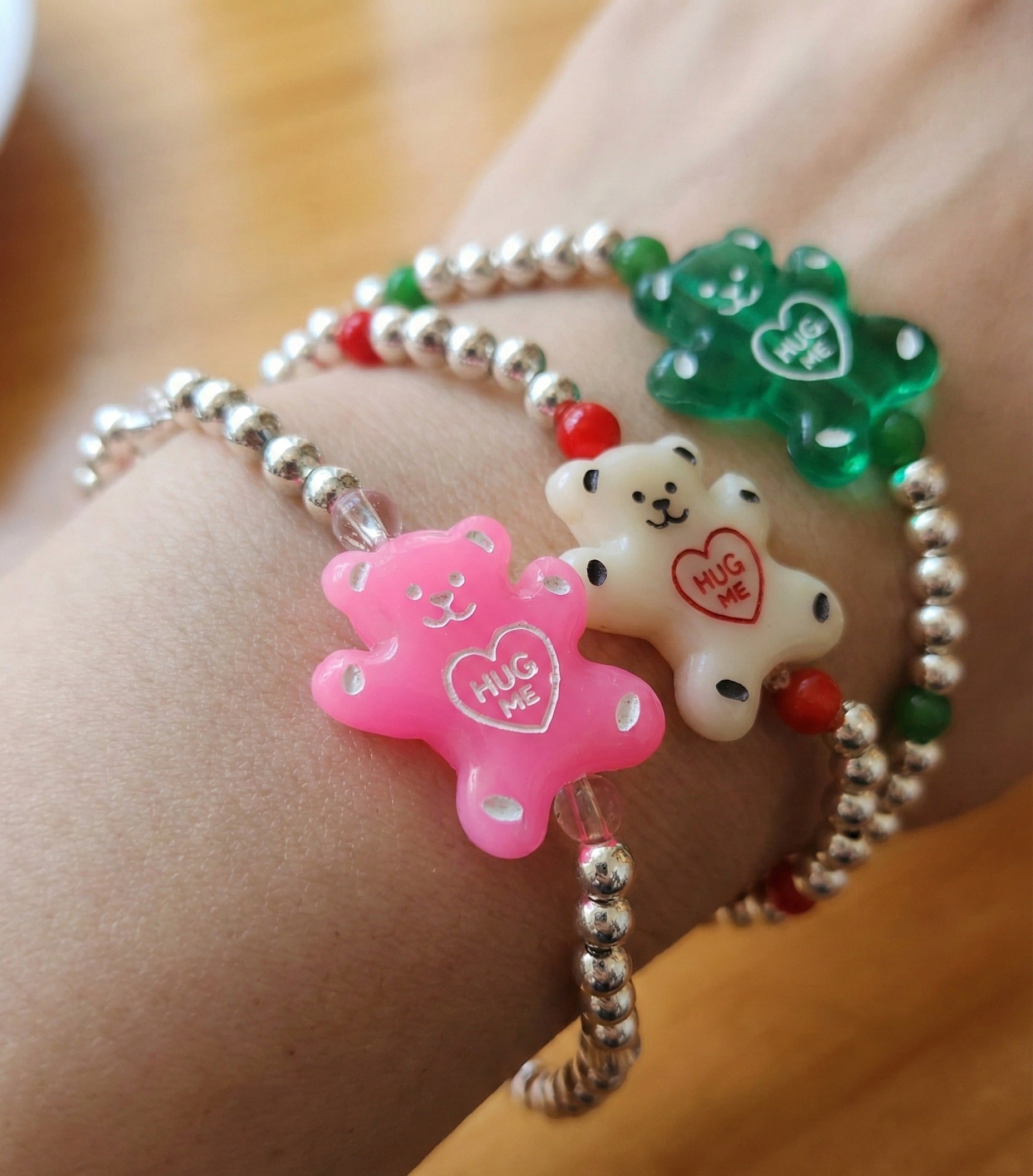 Three Hug Me gummy bear silver ball bracelets — pink, white, and green — overlapping on wooden table with coffee cup