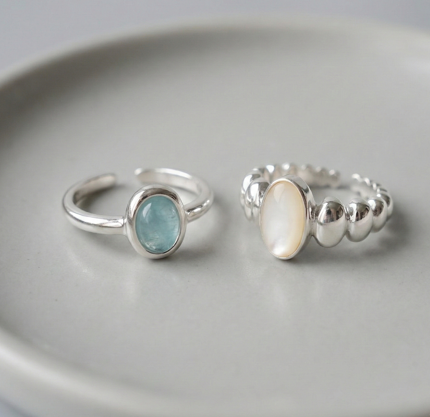 Two 925 sterling silver open rings displayed on white ceramic plate - natural aquamarine ring with smooth band and mother of pearl ring with sculptural ball band side by side