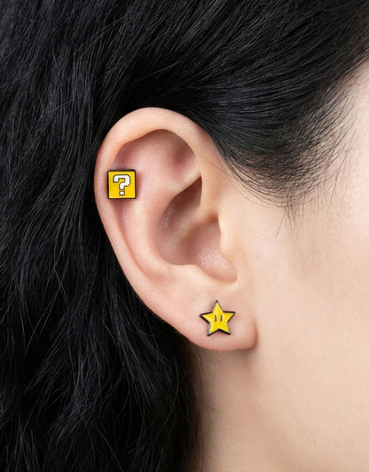 A person wearing mismatched Super Mario-themed earrings: a yellow star stud on the lobe and a yellow question block on the upper ear.
