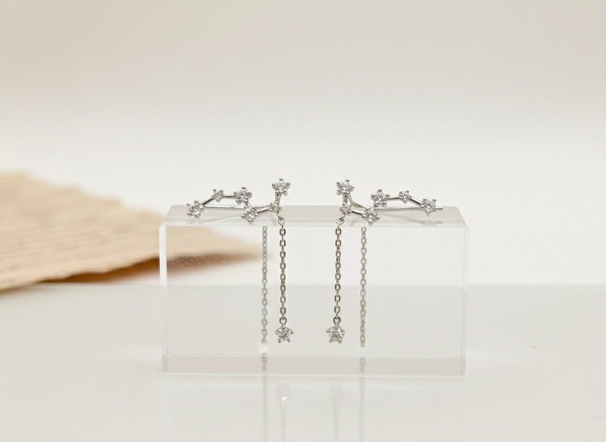 Constellation star earrings with cubic zirconia stones displayed on clear acrylic stand, silver-plated brass with delicate chain drops and 925 sterling silver posts, handcrafted Korean jewelry on neutral beige background
