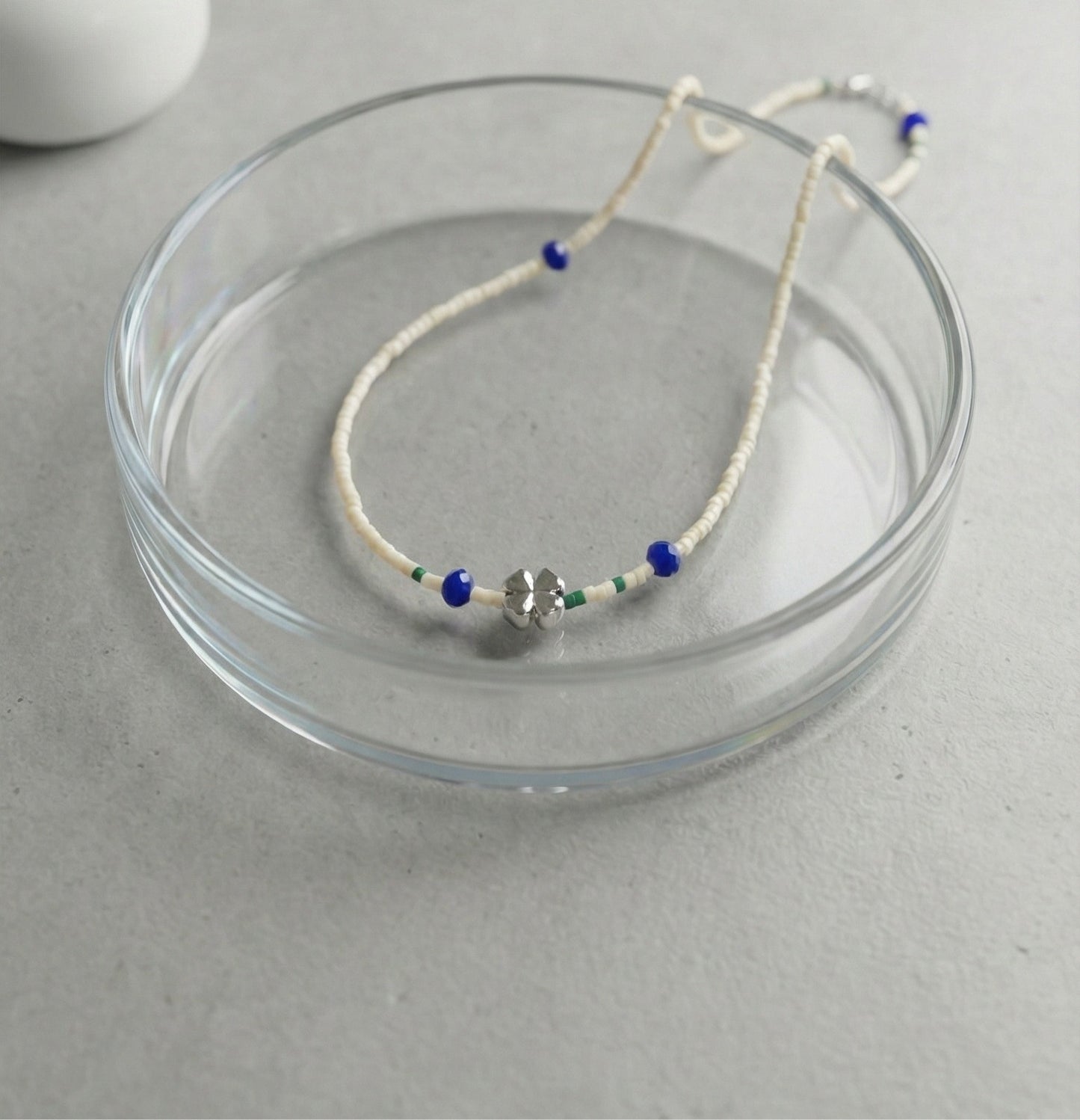 Close-up of ivory seed bead necklace with silver four-leaf clover charm and blue accent beads on glass dish