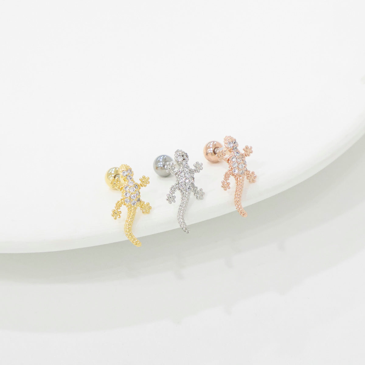 Three gecko-shaped earrings in gold, silver, and rose gold on a white background