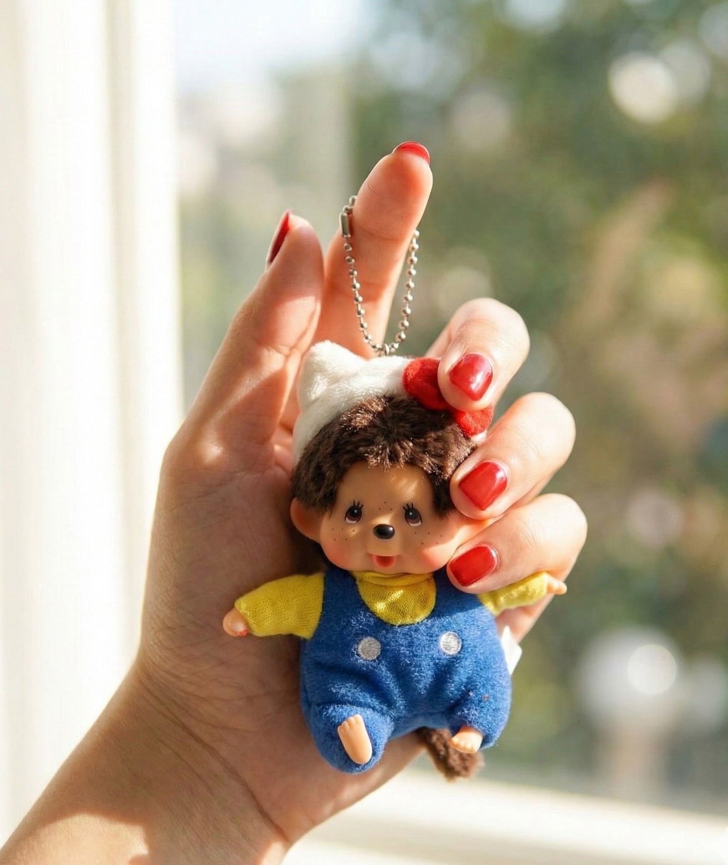Hand holding kawaii Monchhichi Hello Kitty keychain near window with soft natural light