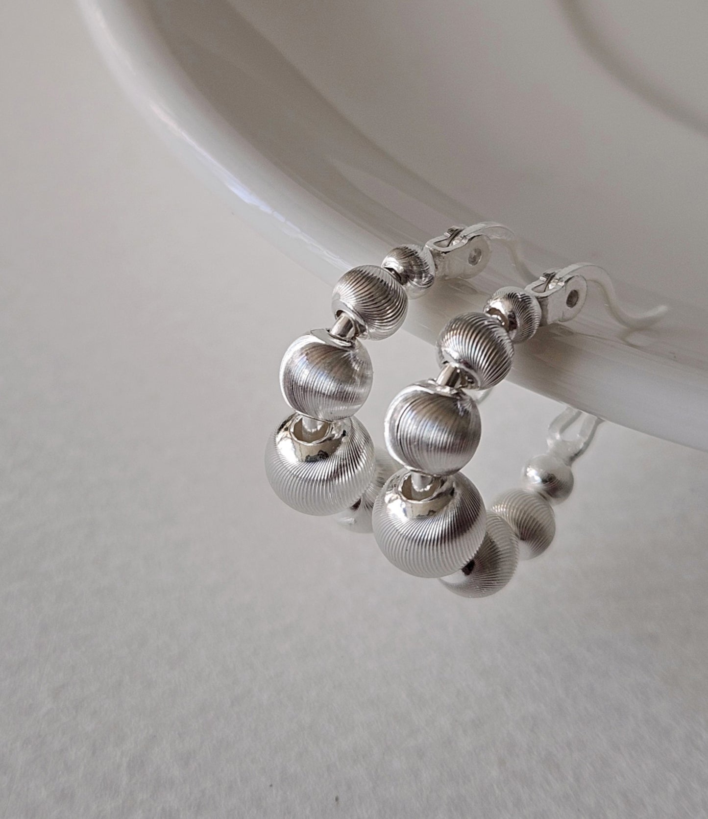 Close-up of silver textured ball hoop earrings on clear acrylic display, showing brushed metal finish with graduated sphere design, 925 sterling silver hypoallergenic posts, minimalist modern jewelry
