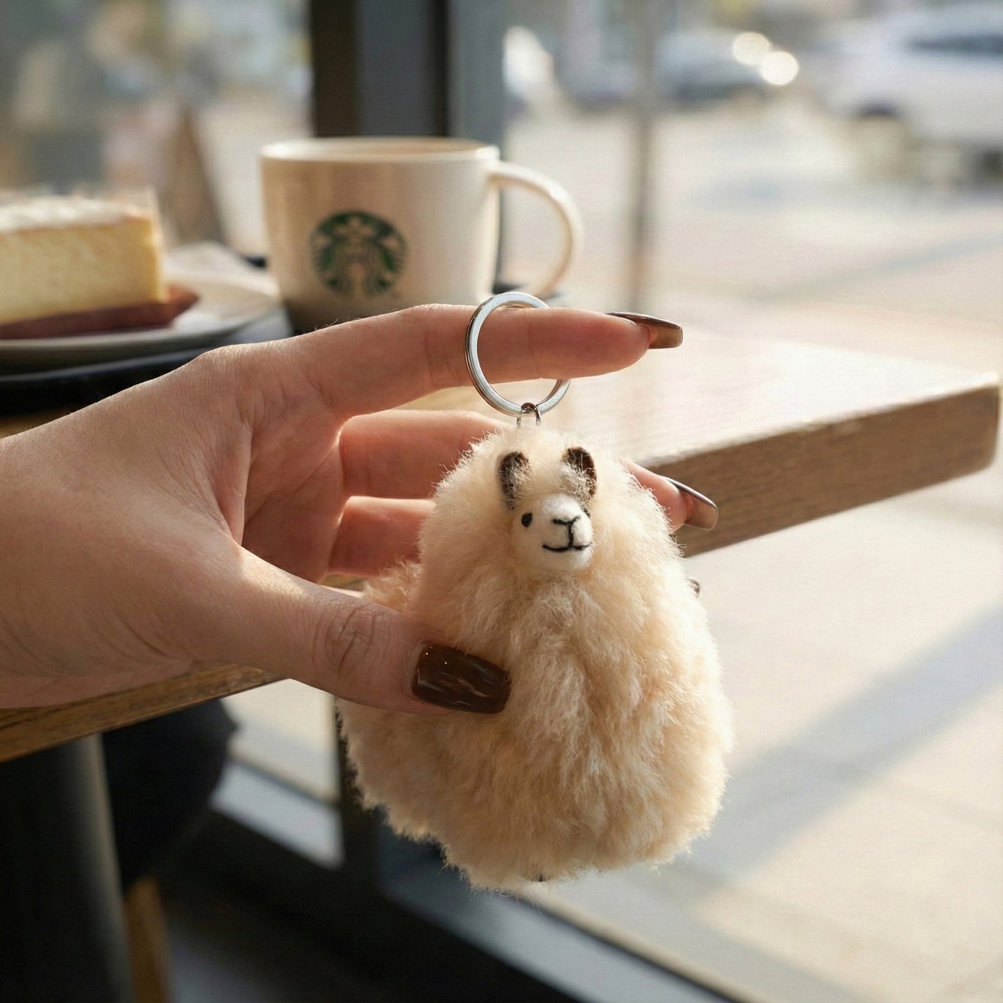 Hand holding beige alpaca fur keychain at a café with coffee cup