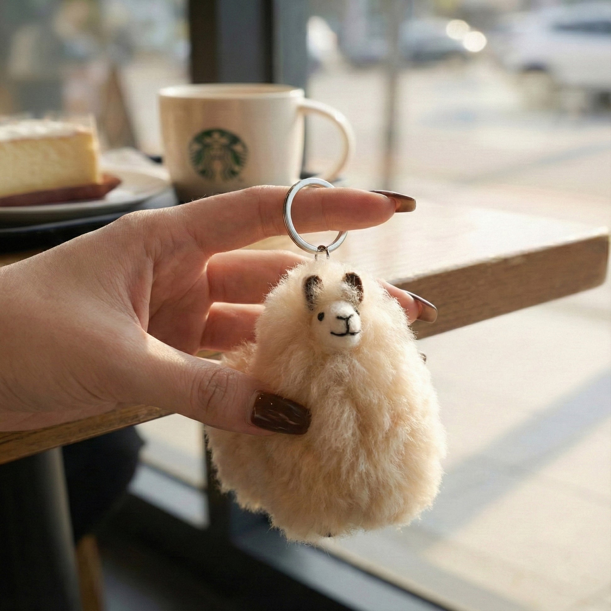 Hand holding beige alpaca fur keychain at a café with coffee cup