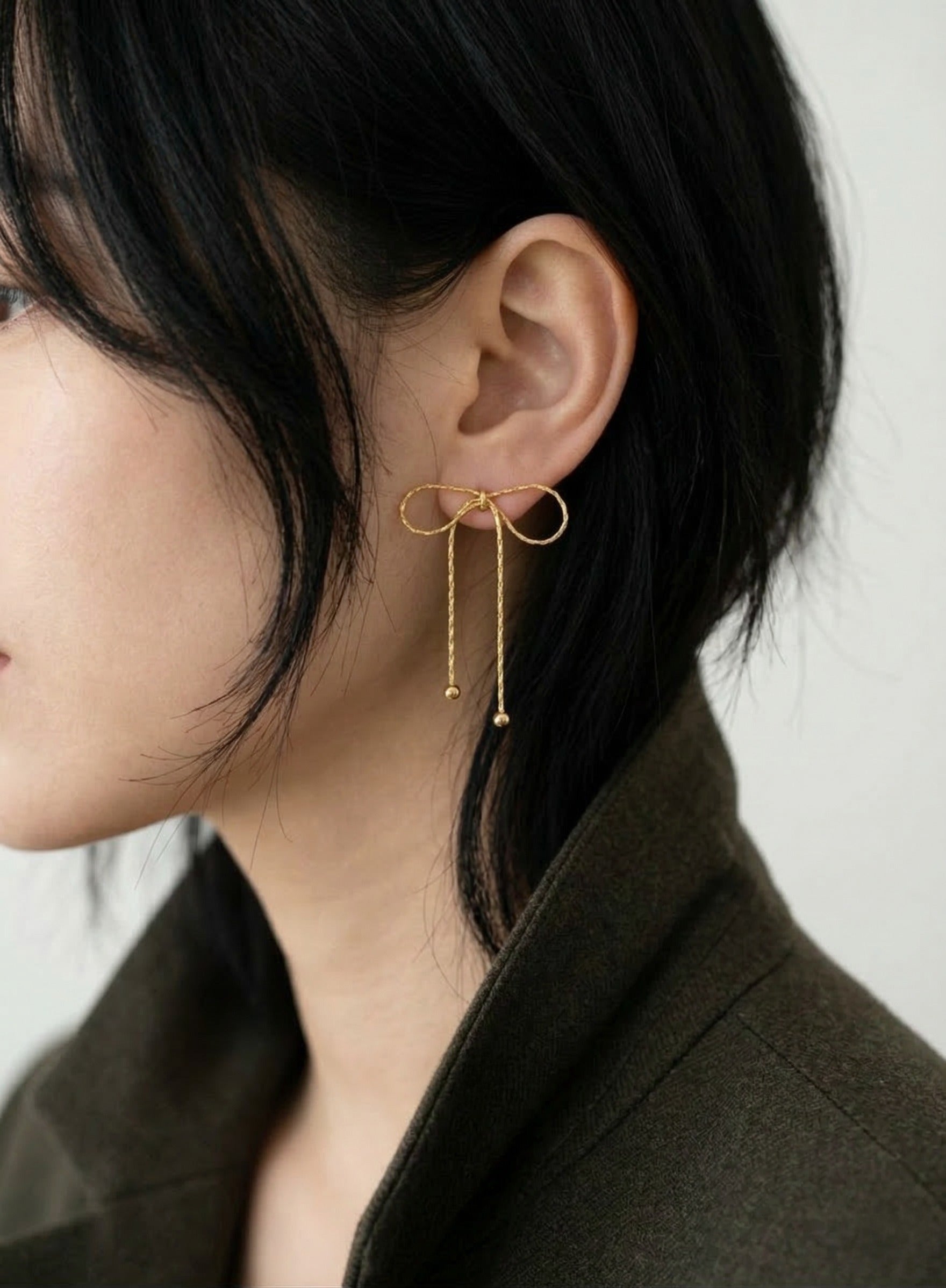 Model wearing gold textured chain bow drop earrings with dark blazer, side profile with hair down, showing full 45mm drop length