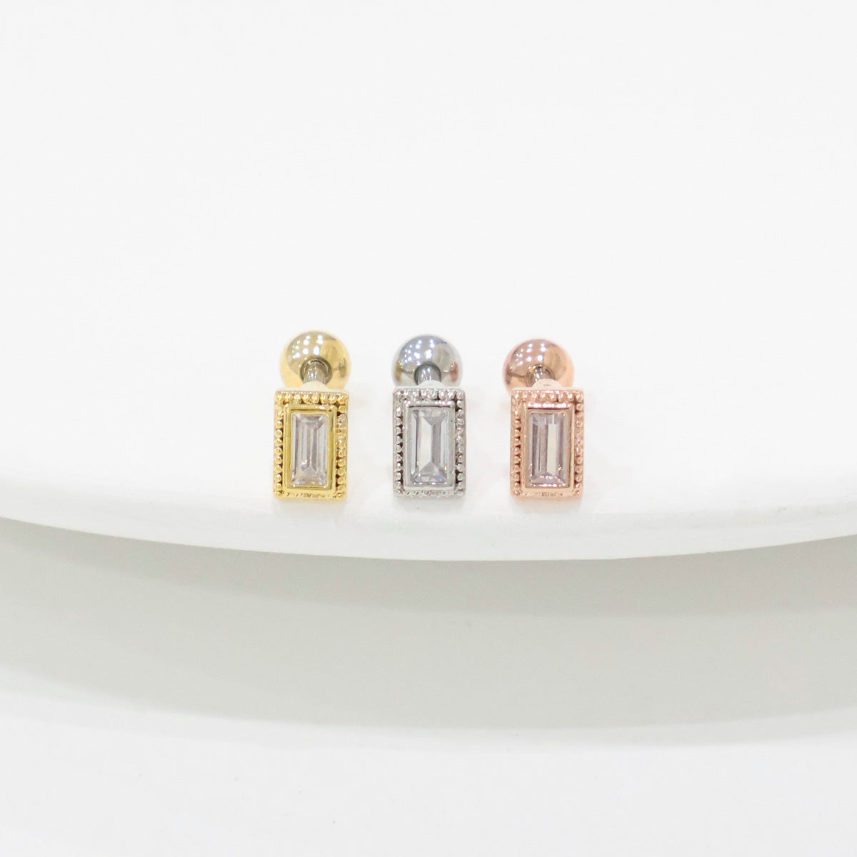 Three rectangular CZ barbell earrings in clear stones, displayed in gold, silver, and rose gold settings on a white curved surface.