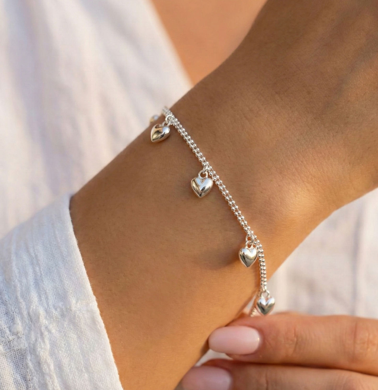 Woman wearing a sterling silver double chain heart charm bracelet on her wrist with a white linen blouse in warm sunlight, showing the puffed heart charms catching light
