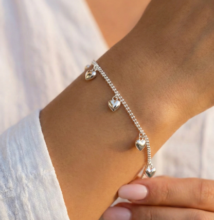 Woman wearing a sterling silver double chain heart charm bracelet on her wrist with a white linen blouse in warm sunlight, showing the puffed heart charms catching light