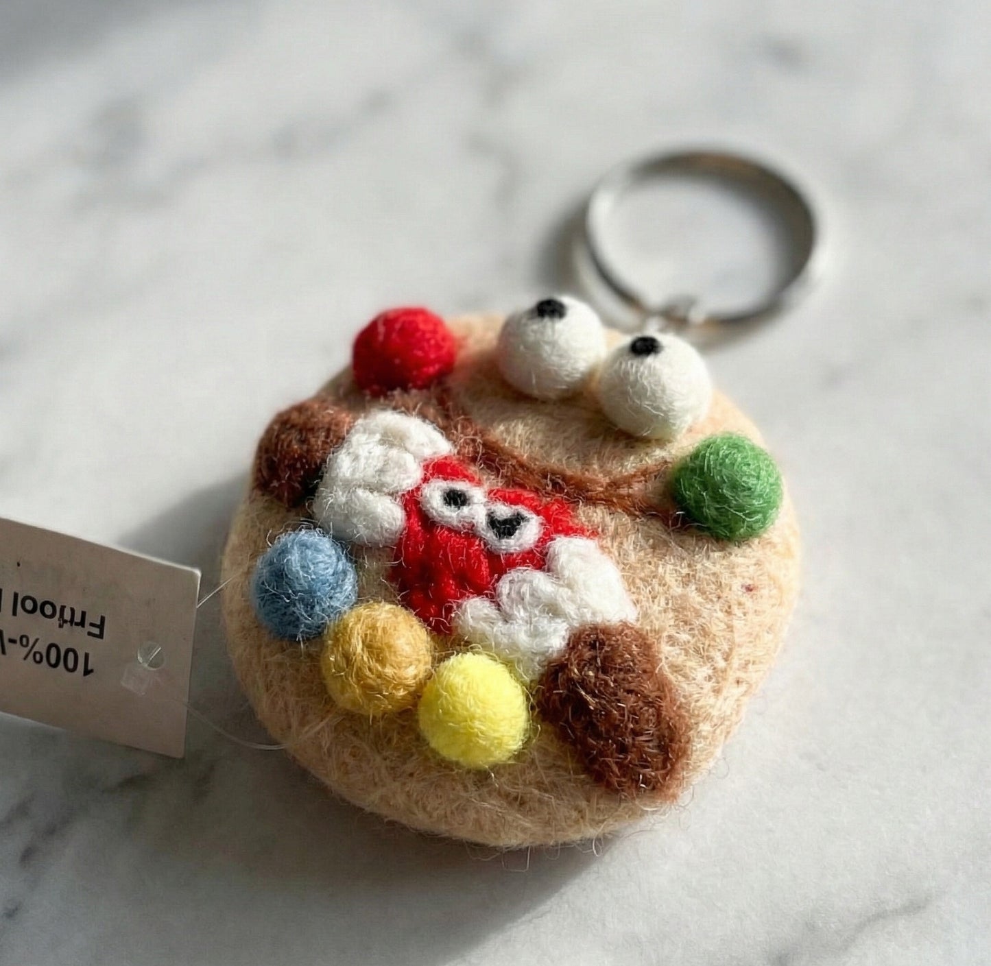 Close-up of cookie monster wool felt keychain with colorful pom-poms and heart detail, handmade in Nepal label visible