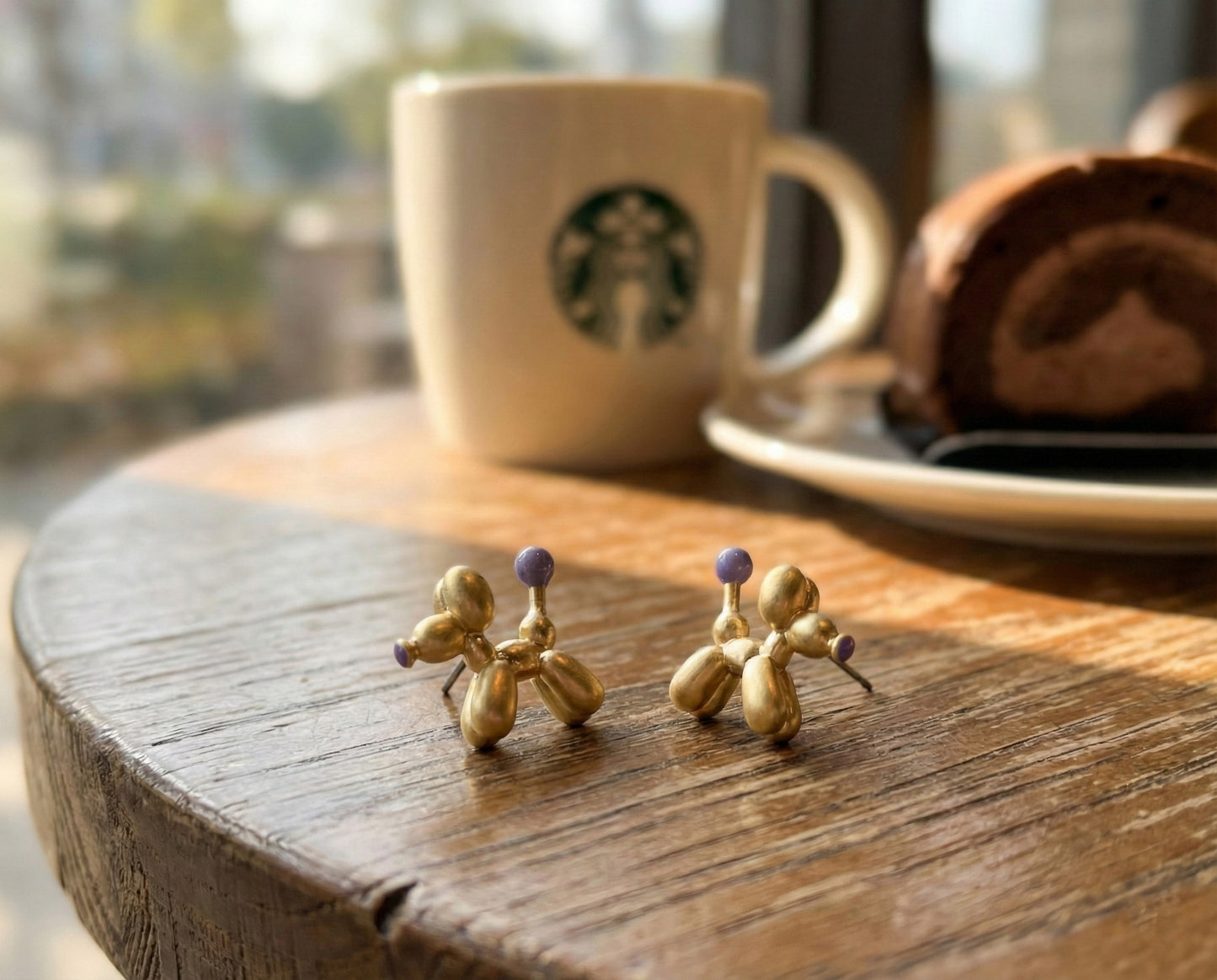 Gold balloon dog earrings with purple accent on wooden tray in natural sunlight showing 3D sculptural details