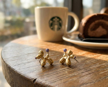 Gold balloon dog earrings with purple accent on wooden tray in natural sunlight showing 3D sculptural details