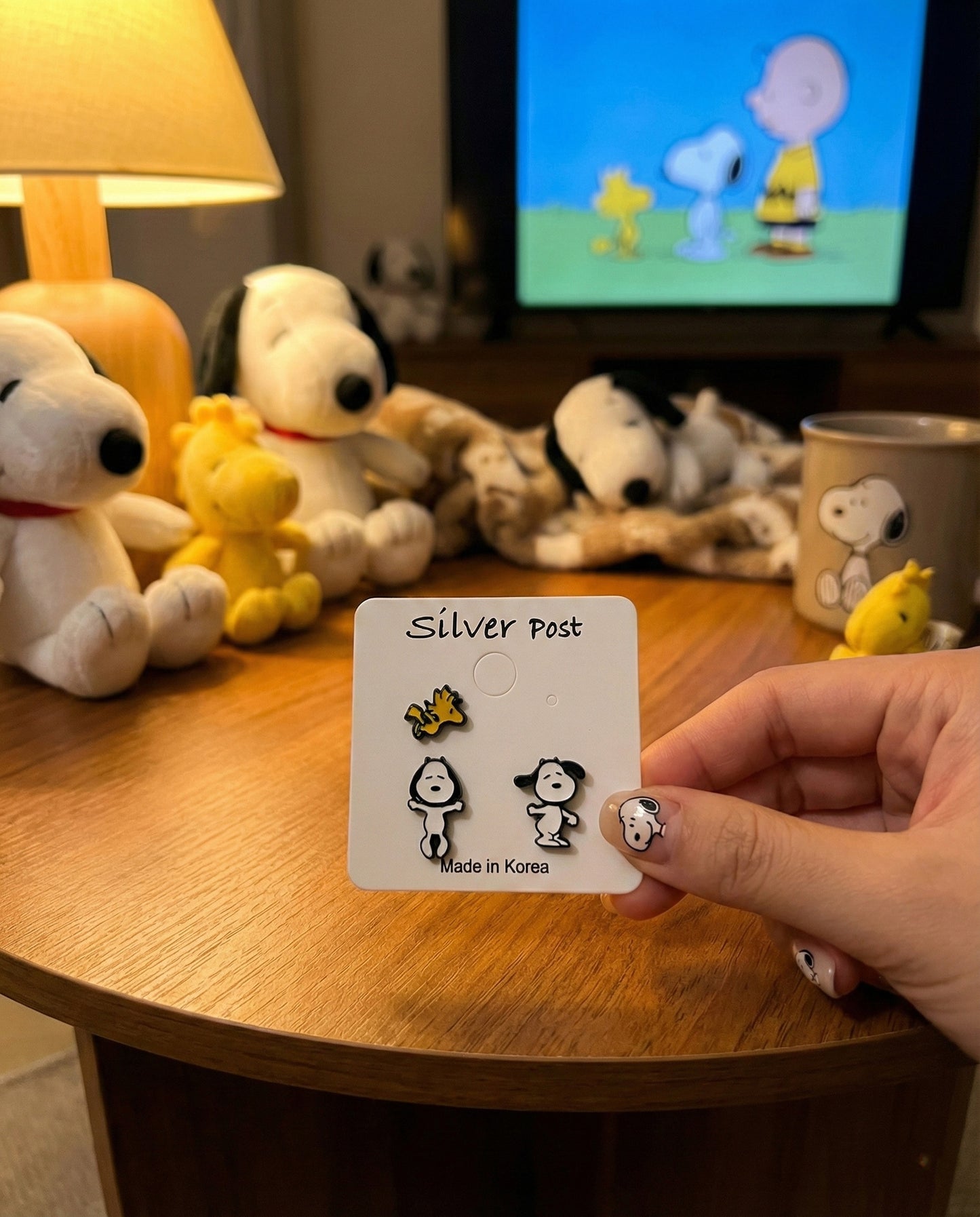 Lifestyle shot of Snoopy and Woodstock 3-piece earring set on card with Peanuts figurines and TV in background
