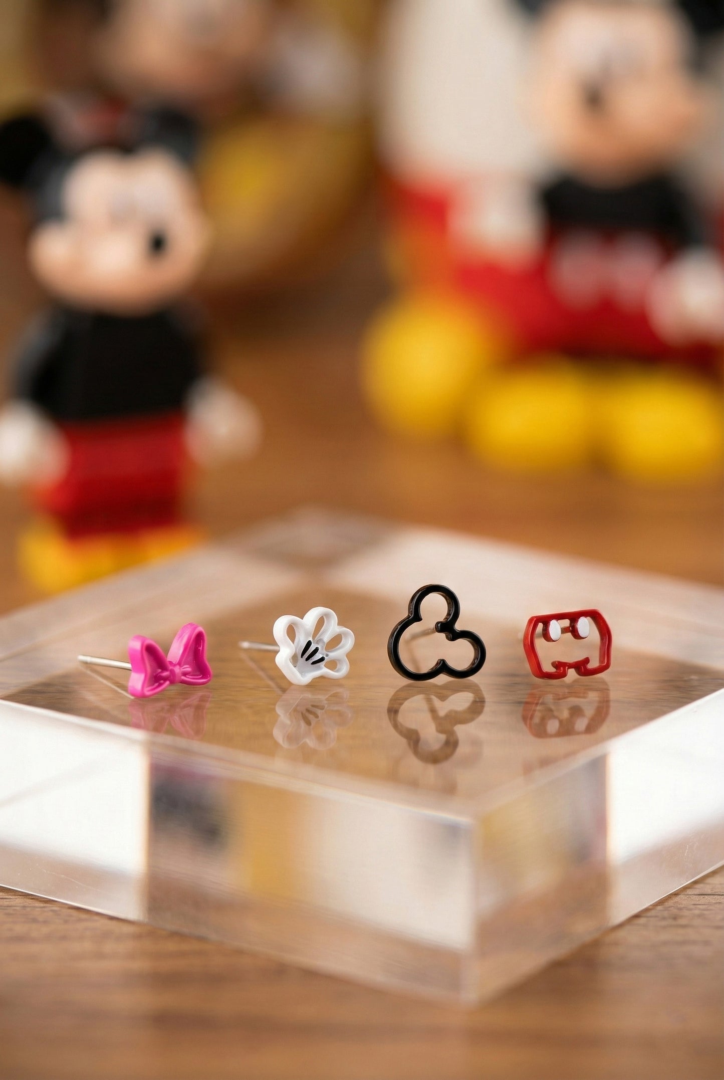 Four Mickey character earrings (pink bow, white glove, black silhouette, red pants) displayed on a clear acrylic block, with Mickey and Minnie figures blurred in the background.