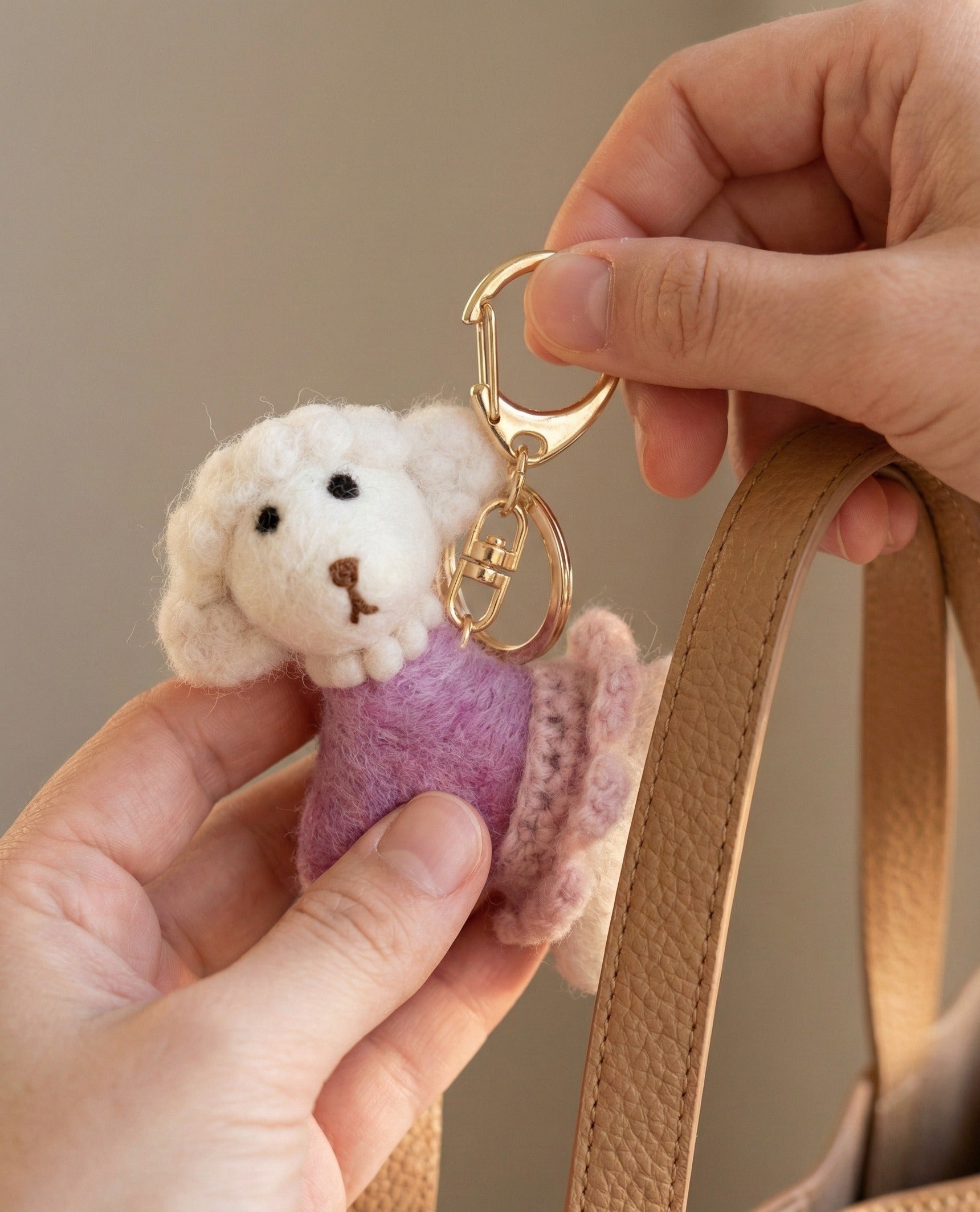 Natural wool poodle keychain with gold hardware attached to tan leather bag strap