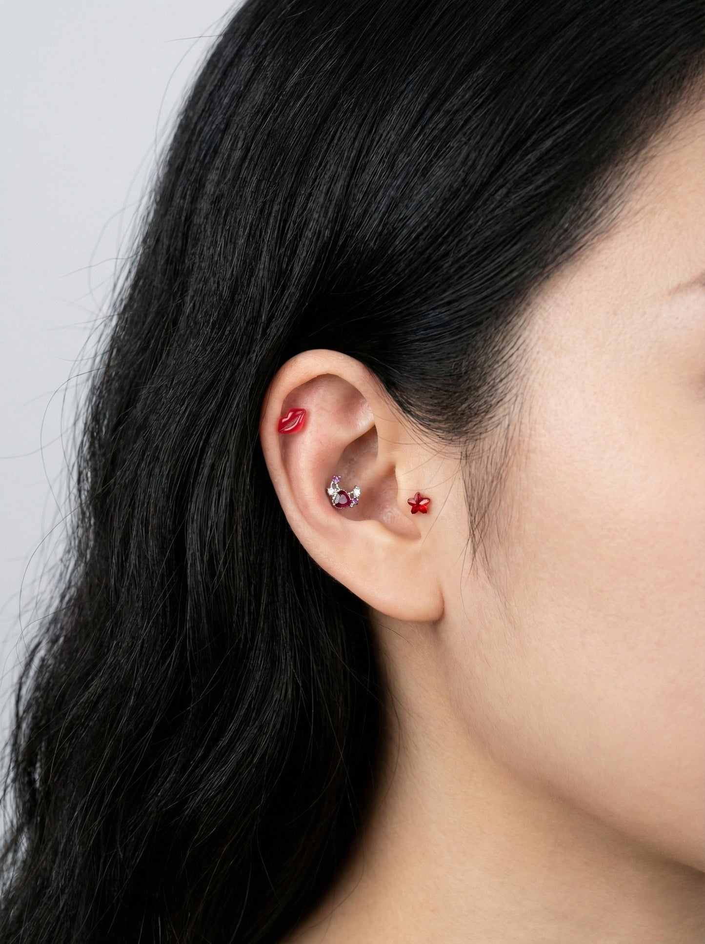 Close-up of red lip-shaped helix piercing and pink moon heart stud on inner conch with red star charm on lobe