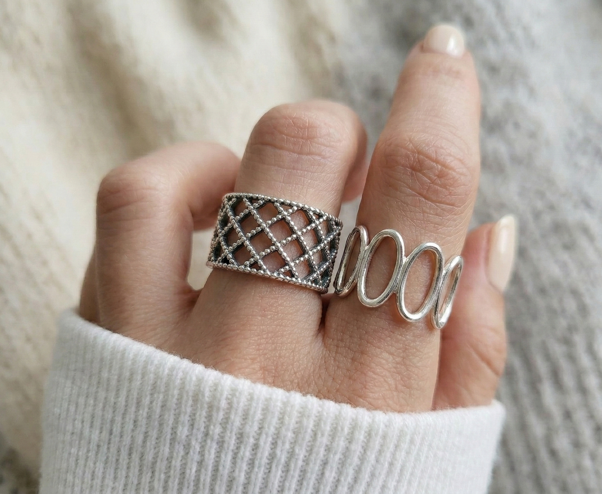 Close-up of lace lattice wide band and oval loop sterling silver rings worn together on hand, white knit background, antique silver finish detail
