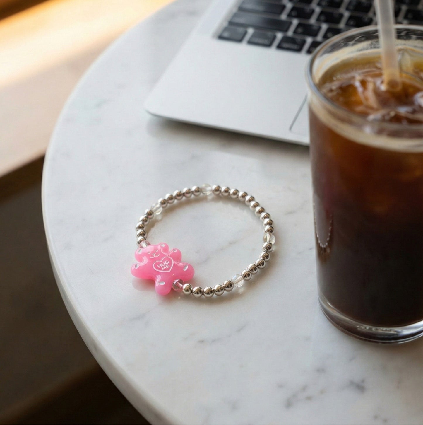 Pink Hug Me gummy bear acrylic charm 925 silver beaded bracelet on marble cafe table next to iced Americano and laptop