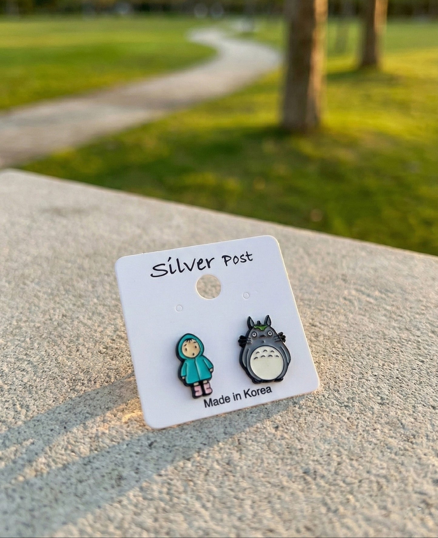 Totoro and Mei earrings on white card labeled Silver Post against green park background