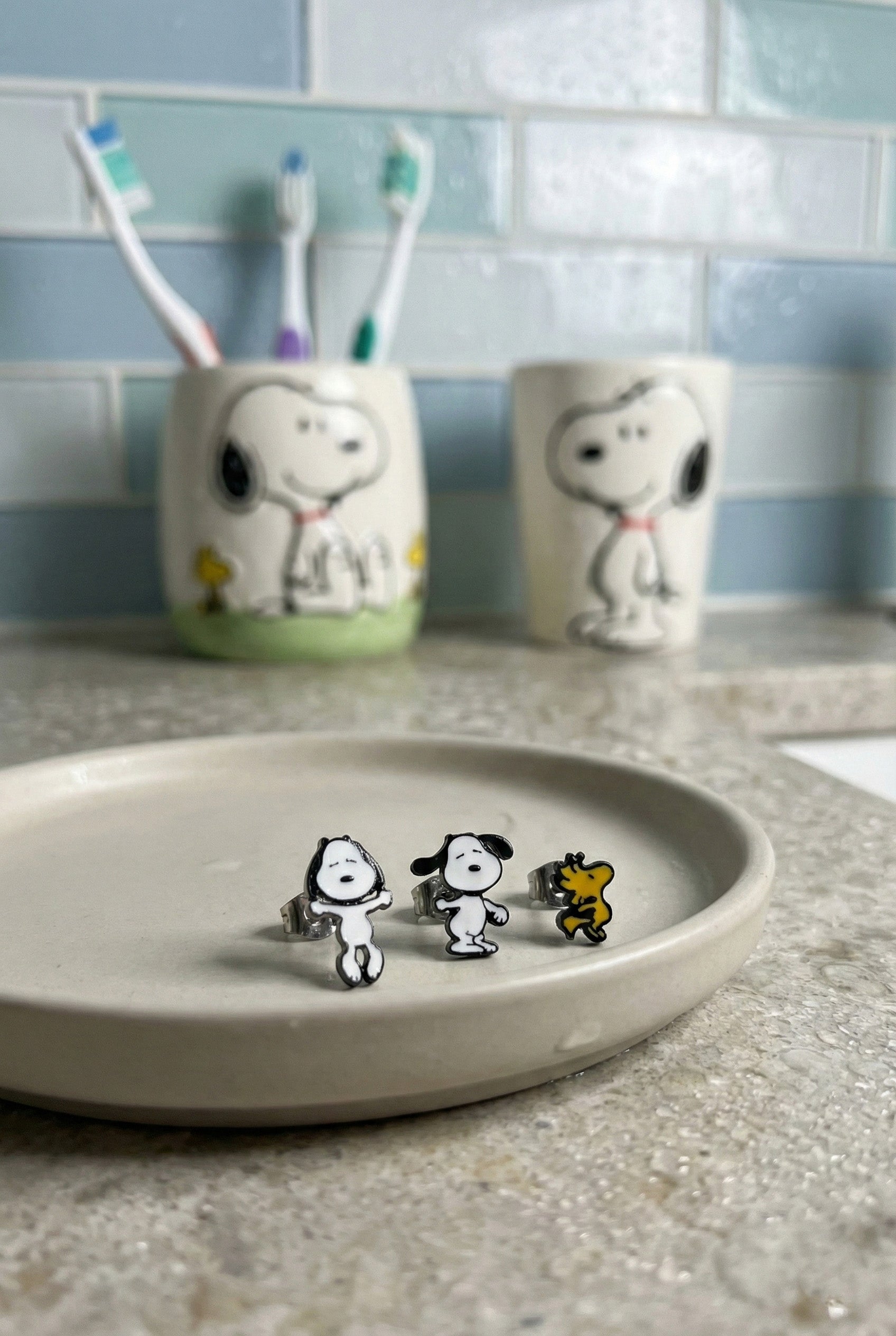 Three piece Snoopy Woodstock earring set displayed on white dish in bathroom setting