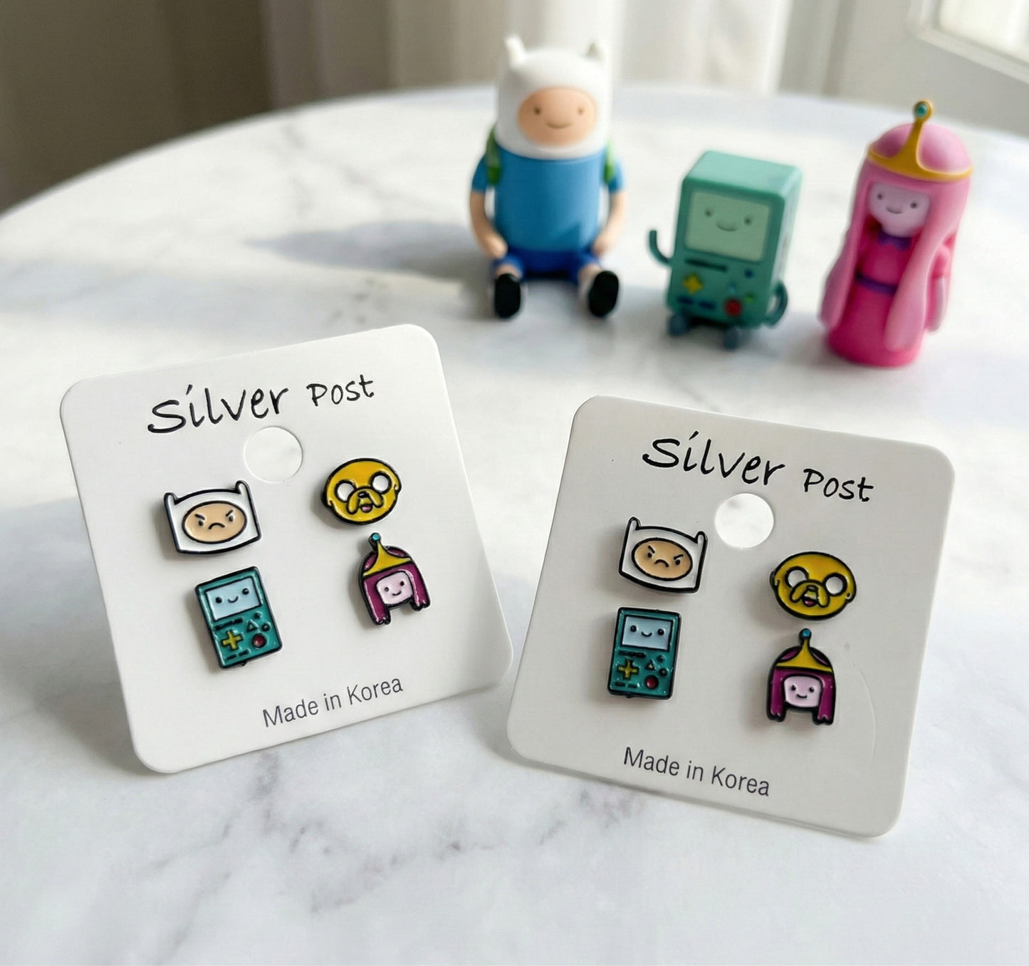 Close-up of Adventure Time enamel earrings Finn angry face Jake BMO Princess Bubblegum with character figurines in background