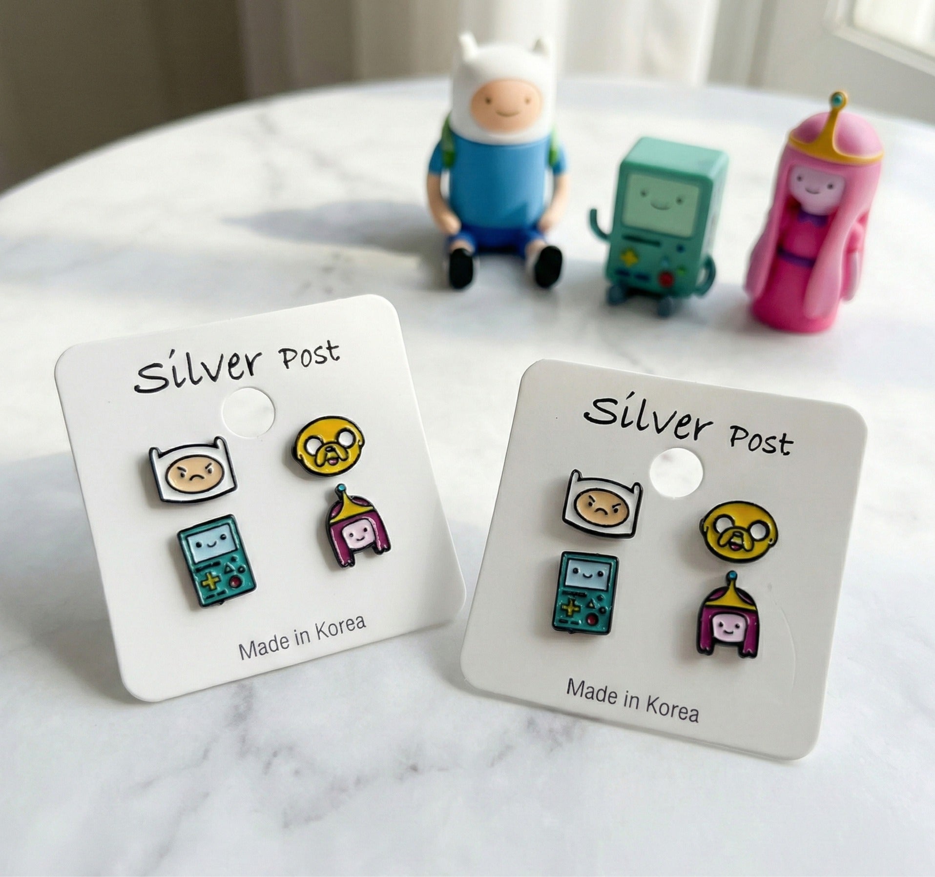 Close-up of Adventure Time enamel earrings Finn angry face Jake BMO Princess Bubblegum with character figurines in background