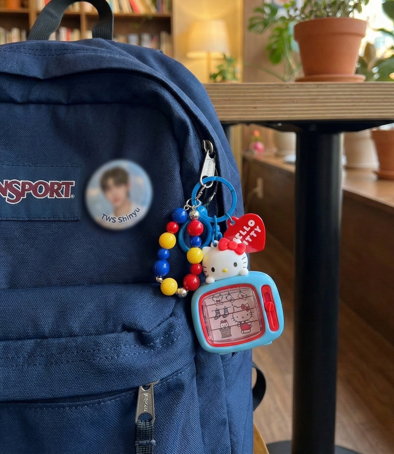  Hello Kitty retro TV bag charm keychain attached to navy blue JanSport backpack with TWS Shinyu pin button in cafe setting — Y2K kawaii school aesthetic